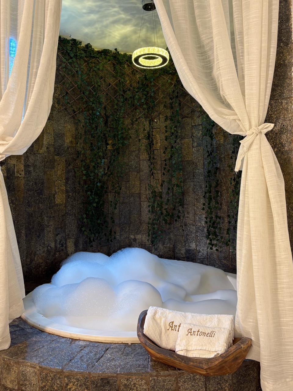 Banheira com espuma em suíte privativa, iluminação suave e cortinas — Studio Antonelli.